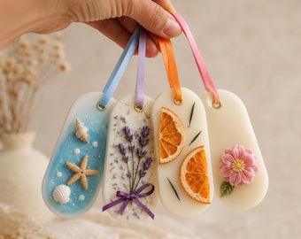 May include: Four oval-shaped wax tablets with ribbons. Each tablet has a different design: a seashell scene, lavender sprigs, orange slices, and a pink flower. The tablets are held by a hand.