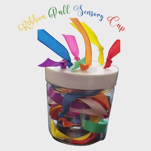 Può includere: Un barattolo di vetro trasparente con coperchio bianco pieno di nastri colorati. I nastri sono di vari colori, tra cui blu, rosa, giallo, arancione, verde e viola. Il testo "Ribbon Pull Sensory Cup" è scritto sopra il barattolo.