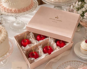 May include: A pink box containing six red cherry-shaped candles with yellow stems, nestled in white paper. The box is labelled "cherry candle" with a cherry logo. Cakes and a cupcake are in the background.