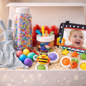 Pode incluir: Uma caixa de presente cheia de brinquedos para bebês, incluindo um coelho de pelúcia, um pote de contas coloridas e uma foto emoldurada de um bebê sorrindo. Também estão incluídos ovos coloridos e vários brinquedos sensoriais.