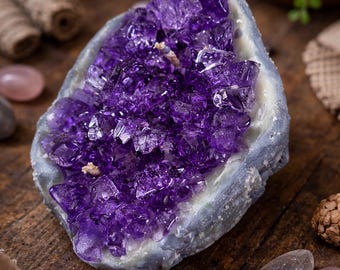 May include: A striking amethyst geode, showcasing a vibrant purple crystal interior within a grey-blue rock. It rests on a wooden surface, accompanied by crystals, a lit candle, and other decorative elements.