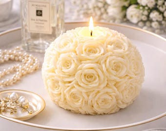 May include: A spherical candle with a rose design, lit with a single flame. The candle is ivory-coloured and sits on a white tray with gold trim. Other items include a perfume bottle, jewellery, and flowers.