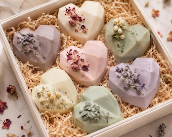 May include: A white gift box containing seven heart-shaped soaps in pastel shades. Each soap is adorned with dried flowers and herbs, such as lavender and rose petals, and sits on a bed of shredded paper.