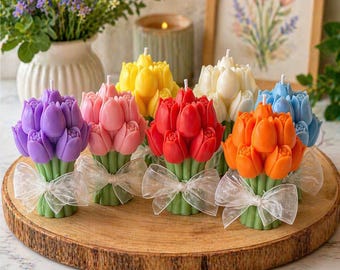 May include: A selection of multi-coloured tulip-shaped candles, including purple, pink, red, yellow, orange, white, and blue. Each candle is tied with a white ribbon. The candles are displayed on a wooden surface.