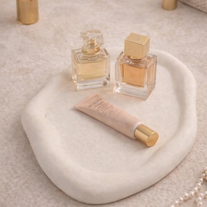 May include: A collection of beauty products on a white, organic-shaped tray. Includes two perfume bottles with gold caps, a tube of Beauty Cream, and a gold makeup brush. The products are arranged on a neutral-toned surface.