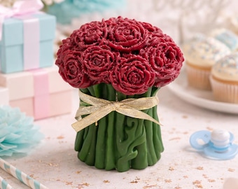 May include: A burgundy rose bouquet candle with green stems, tied with a gold ribbon. The candle is surrounded by baby shower decorations, including cupcakes, a dummy, and gift boxes. The word "baby" is visible in the background.