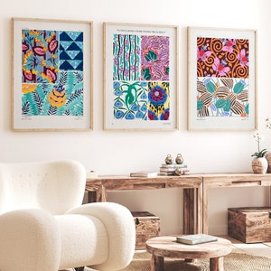 Könnte beinhalten: Drei gerahmte Drucke mit bunten abstrakten Mustern mit floralen und geometrischen Designs. Die Drucke hängen an einer weißen Wand über einem Holztisch und einem weißen Sessel.