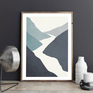 Op de afbeelding: Abstracte kunstprint met een witte rivier die door een blauw en grijs berglandschap slingert. De print is ingelijst in een licht houten lijst.