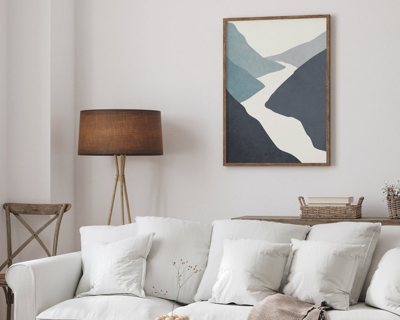Op de afbeelding: Een witte bank met witte kussens en een bruine deken. Een bruine staande lamp met een bruine lampenkap staat op de achtergrond. Een ingelijst afdruk van een blauw en wit abstract landschap hangt aan de muur boven de bank.