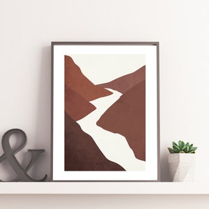 Op de afbeelding: Abstracte kunstprint met een minimalistisch landschap met een rivier die door bruine bergen slingert. De print is ingelijst in een zwart frame en wordt getoond op een witte plank.
