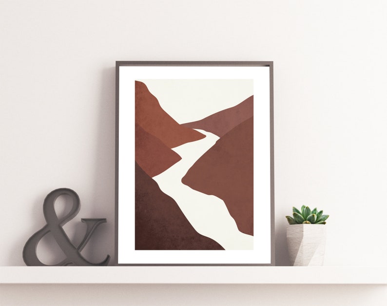 Op de afbeelding: Abstracte kunstprint met een minimalistisch landschap met een rivier die door bruine bergen slingert. De print is ingelijst in een zwart frame en wordt getoond op een witte plank.