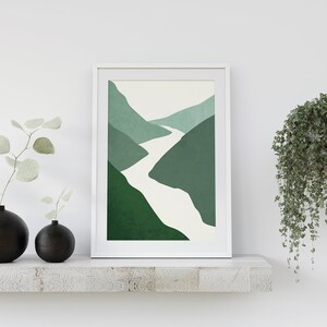 Op de afbeelding: Een ingelijst abstract print van een witte rivier die door groene bergen slingert. De print hangt aan een witte muur boven een plank met twee zwarte vazen en een hangplant.