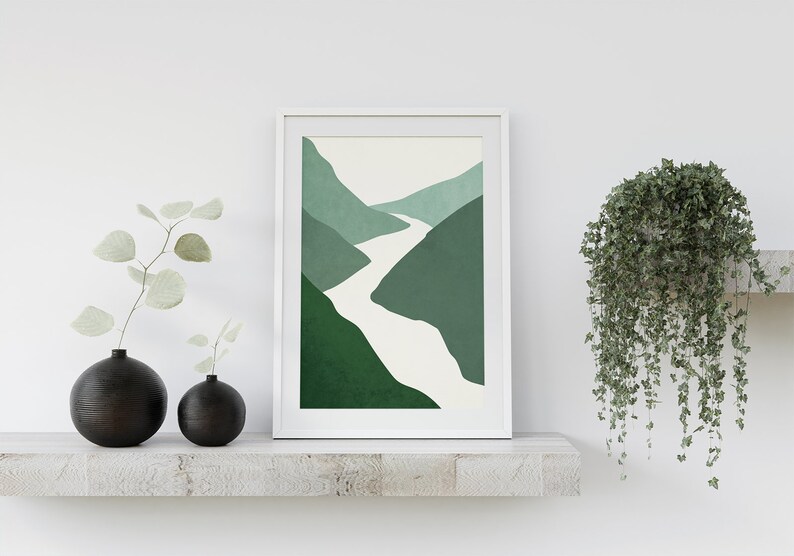 Op de afbeelding: Een ingelijst abstract print van een witte rivier die door groene bergen slingert. De print hangt aan een witte muur boven een plank met twee zwarte vazen en een hangplant.