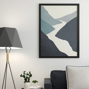 Op de afbeelding: Een zwarte ingelijste abstracte print van een witte rivier die door blauwe en grijze bergen slingert. De print hangt aan een witte muur boven een zwarte bank met een wit kussen.
