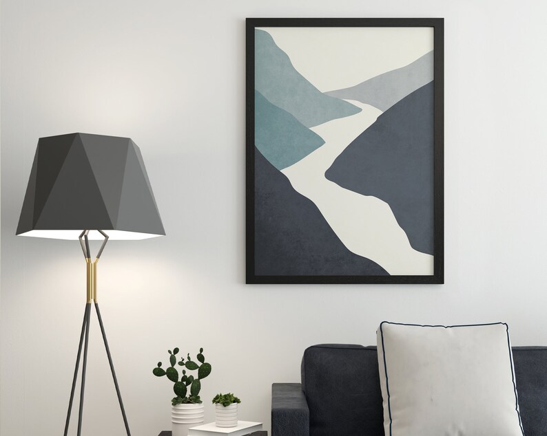Op de afbeelding: Een zwarte ingelijste abstracte print van een witte rivier die door blauwe en grijze bergen slingert. De print hangt aan een witte muur boven een zwarte bank met een wit kussen.