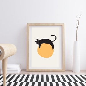 Puede incluir: Una impresi&oacute;n enmarcada de un gato negro acostado sobre un c&iacute;rculo amarillo. El gato tiene ojos grandes y una cola enroscada. El fondo es de color crema claro.