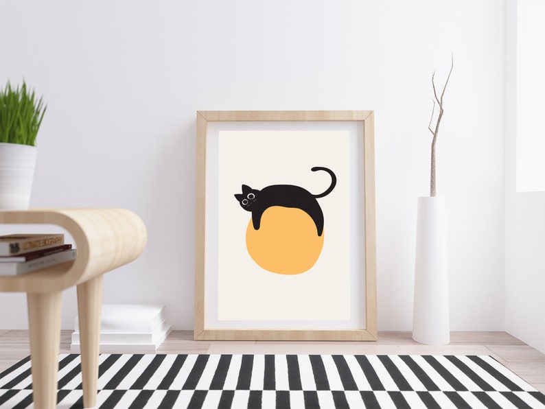 Puede incluir: Una impresi&oacute;n enmarcada de un gato negro acostado sobre un c&iacute;rculo amarillo. El gato tiene ojos grandes y una cola enroscada. El fondo es de color crema claro.