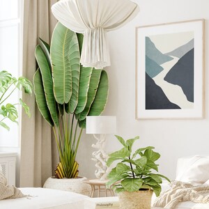Op de afbeelding: Een witte bank met een beige deken en witte kussens in een woonkamer met een grote groene plant, een kleinere groene plant in een geweven mand, een witte lamp en een ingelijst abstract drukwerk van een blauw en wit landschap.