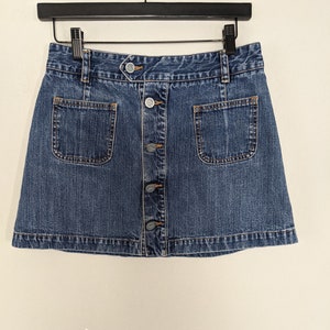 May include: A blue denim skirt with a button-up front and two front pockets. The skirt is hanging on a black hanger against a white background. The skirt has a classic, casual style.