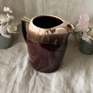 Puede incluir: Una jarra de cerámica esmaltada marrón y blanca con una boca ancha y un asa. La jarra tiene un esmalte marrón con una banda blanca alrededor de la parte superior.