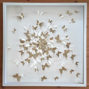 May include: A white framed square wall art piece with a gold and white butterfly design. The butterflies are arranged in a circular pattern with the center being the densest.