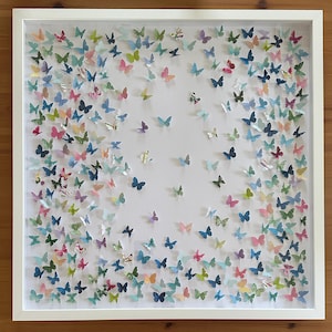May include: A white-framed artwork filled with a multitude of paper butterflies in various colours. The butterflies, in shades of blue, pink, green, and yellow, are set against a white backdrop. The frame's white colour gives a clean, contemporary look.