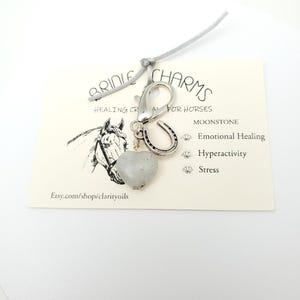 May include: A silver horse bridle charm with a moonstone heart, horseshoe, and a silver clip. The charm is attached to a gray cord. The card reads "Bridle Charms Healing Crystals for Horses Moonstone Emotional Healing Hyperactivity Stress Etsy.com/shop/clarityoils"