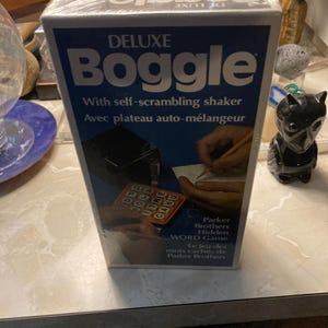 May include: A Deluxe Boggle game box with a blue and white design. The box features the game's name in large letters and includes the text "With self-scrambling shaker". The box also shows images of the game in use.
