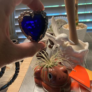 May include: A heart-shaped brooch featuring a large, deep blue faceted center stone surrounded by a border of clear, sparkling rhinestones set in a silver-toned metal frame, held up to display the vibrant color and reflective surface.