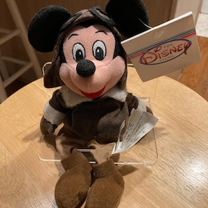 May include: A Mickey Mouse plush toy wearing a brown aviator hat and jacket. The plush has black ears, a pink face, and brown shoes. A tag from The Disney Store is attached.