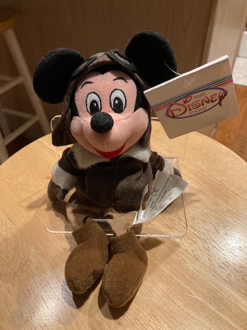 May include: A Mickey Mouse plush toy wearing a brown aviator hat and jacket. The plush has black ears, a pink face, and brown shoes. A tag from The Disney Store is attached.