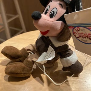 May include: A plush Mickey Mouse toy dressed as a pilot, wearing a brown aviator cap with goggles and a brown jacket with white trim. The toy has a pink face, black nose, and red mouth. A tag from The Disney Store is visible.
