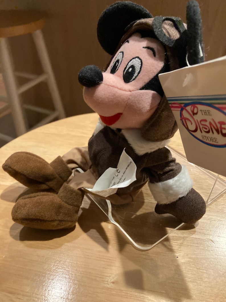 May include: A plush Mickey Mouse toy dressed as a pilot, wearing a brown aviator cap with goggles and a brown jacket with white trim. The toy has a pink face, black nose, and red mouth. A tag from The Disney Store is visible.