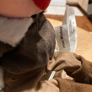 May include: A plush toy of Mickey Mouse dressed as a pilot. The toy has a brown jacket, tan pants, and a white scarf. The tag reads "Pilot Mickey". The toy is made in China.