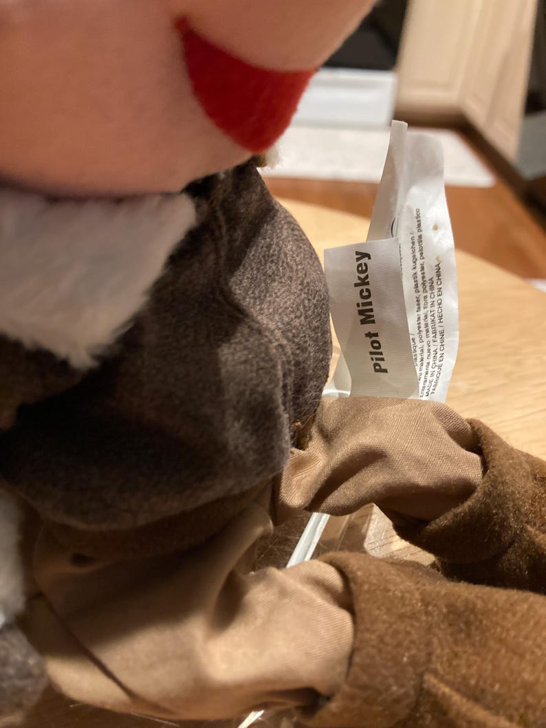May include: A plush toy of Mickey Mouse dressed as a pilot. The toy has a brown jacket, tan pants, and a white scarf. The tag reads "Pilot Mickey". The toy is made in China.