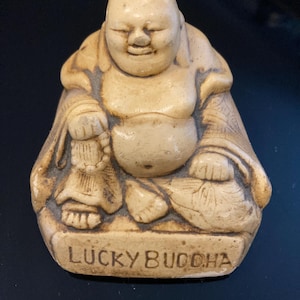 May include: A beige-colored Lucky Buddha figurine. The seated figure has a large belly, closed eyes, and a serene expression. The base of the figurine is inscribed with the words "LUCKY BUDDHA". The figurine is made of a stone-like material.