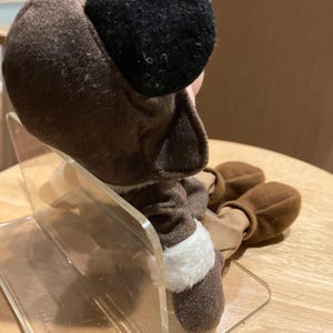 May include: A brown plush toy with black ears and a white trim. The toy is sitting on a clear acrylic stand. The toy's feet are also brown.