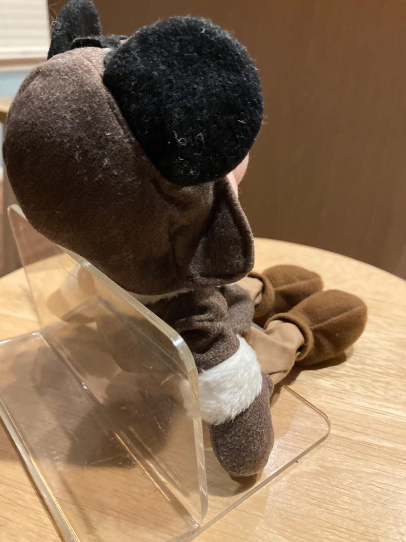 May include: A brown plush toy with black ears and a white trim. The toy is sitting on a clear acrylic stand. The toy's feet are also brown.