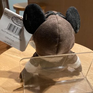 May include: A brown Pilot Mickey Mouse bean bag plush toy with black ears and a pilot's cap. The plush is wearing a white and brown outfit. A Disney Store tag is attached.
