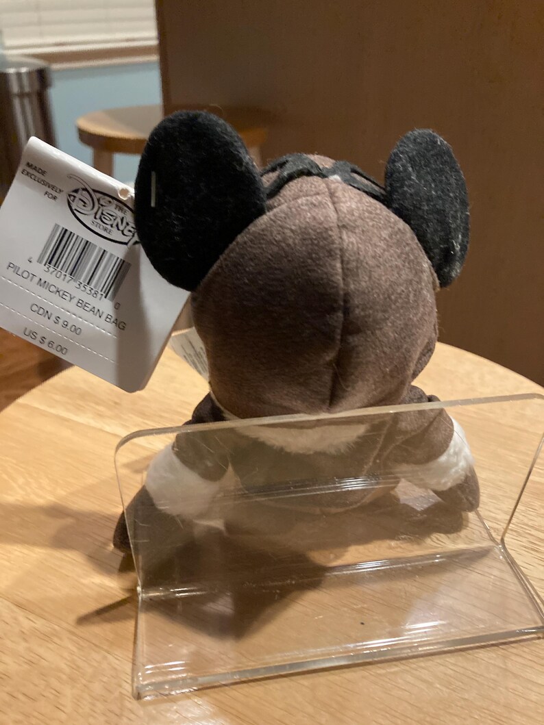 May include: A brown Pilot Mickey Mouse bean bag plush toy with black ears and a pilot's cap. The plush is wearing a white and brown outfit. A Disney Store tag is attached.