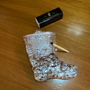 May include: A clear, textured glass boot-shaped ashtray with a match inserted. A black box of matches with the text "J.C. Newman Cigar Co." is next to the ashtray. The items are on a wooden surface.