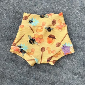 May include: Yellow baby shorts with a repeating pattern of bees, honeycombs, honey pots, and honey dippers. The shorts have a high waist and a textured fabric. The overall design has a whimsical, playful theme.
