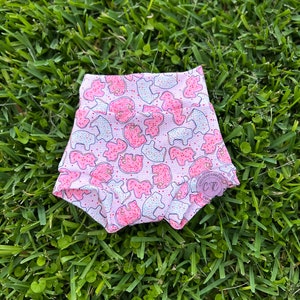 May include: Pink baby shorts with a repeating pattern of pink and white frosted animal cookies with red sprinkles. The shorts are made of a soft, stretchy fabric and are set against a background of green grass.