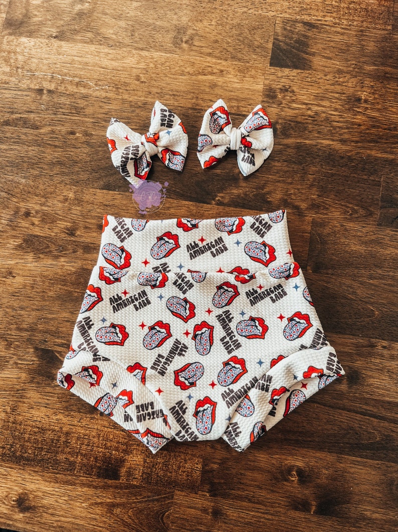 May include: White baby shorts and two matching hair bows. The shorts and bows feature a repeating pattern of red lips, blue stars, and the text "ALL AMERICAN BABE" in black. The fabric has a textured appearance.