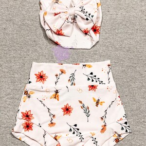 May include: A baby outfit set featuring a headband with a large bow and high-waisted shorts. Both pieces are light pink and adorned with a floral and bee pattern in orange, yellow, and green. The shorts have a rolled hem.