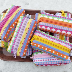 Könnte beinhalten: Eine Sammlung bunter Federmäppchen mit verschiedenen Mustern und Reißverschlüssen. Die Etuis weisen leuchtende Farben wie Pink, Orange, Gelb und Blau mit geometrischen und gepunkteten Designs auf. Sie sind auf einem Holztablett angeordnet.