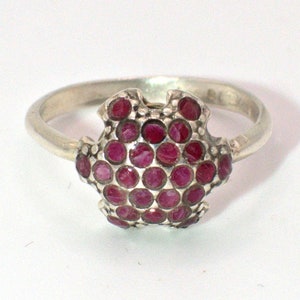 May include: A silver ring with a cluster of small, round, deep red gemstones. The gemstones are set in an ornate, openwork design. The ring has a plain, polished band.