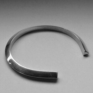 May include: A silver cuff bracelet with a minimalist design. The bracelet has a smooth, curved shape with squared-off ends. The open design allows for easy wear. The metal has a polished finish, reflecting light.