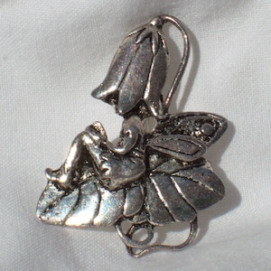 May include: Silver-toned fairy figurine brooch with wings, sitting on a flower with a bell-shaped flower on top.