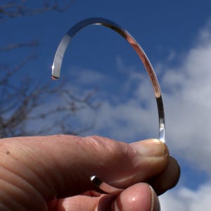 May include: A silver bangle bracelet with a smooth, polished finish. The bracelet is open and has a slight curve. The inscription "925 STERLING" is visible on the inside of the bracelet.